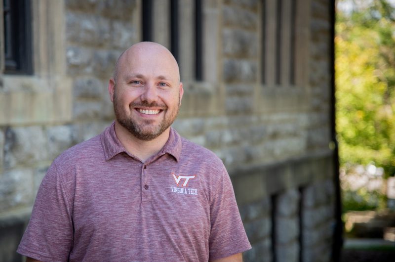 A man, wearing a light maroon Virginia Tech polo shirt, poses by a building comprised of Hokie Stone.