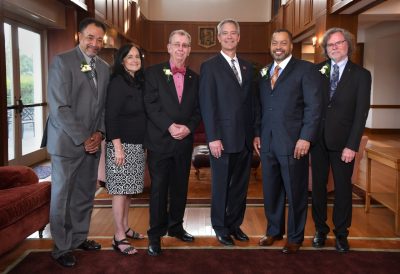 Five men and a woman pose in dress attire  inside a wood-paneled alumni welcome lounge.