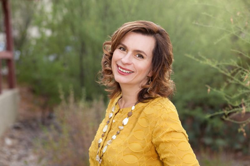 A woman with short brown hair  and a yellow blouse smiles.