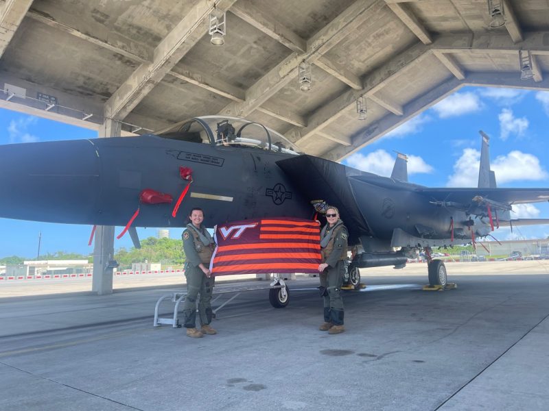 Jacobson and Archer stand holding a striped VT flag in front of a F-15E military fighter jet in a hanger. Both are smiling and wearing flight suits. 
