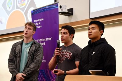 Three students stand at front of classroom with projection screen in background. Middle student holds microphone as he is speaking.