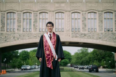 Student in graduation regalia stands with Torgerson Bridge in background.