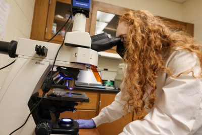 young woman with protective lab equipment looks into microscope