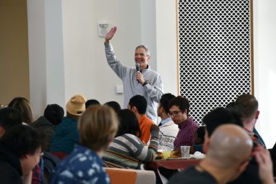 Smiling man with microphone waves to room full of eating students.