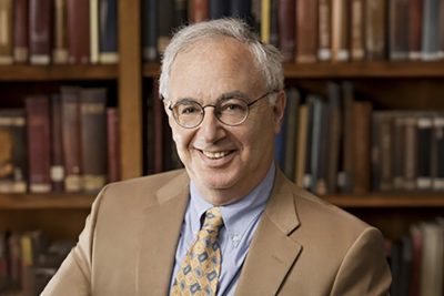 A man is sitting in front of a bookshelf. He is wearing glasses and a tan suit with a blue shirt and yellow and blue tie.