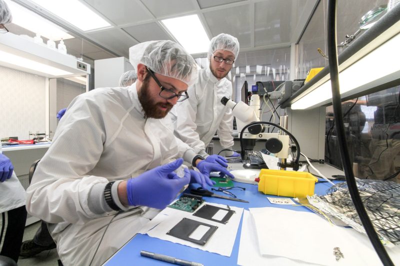 From right to left, Nick Angle and Josh Smoot at lab bench in coats, hair nets, and nitrile gloves, make minor modifications on the custom-made flight board for the CubeSat