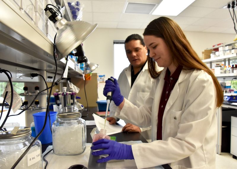 Taylor in foreground pipetting a liquid with Bowers looking over in background. Both wear lab coats and stand at lab bench.