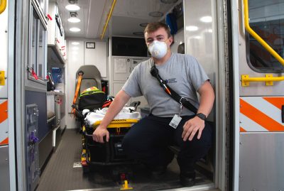 Buck wears a mask and kneels next to gurney inside ambulance.