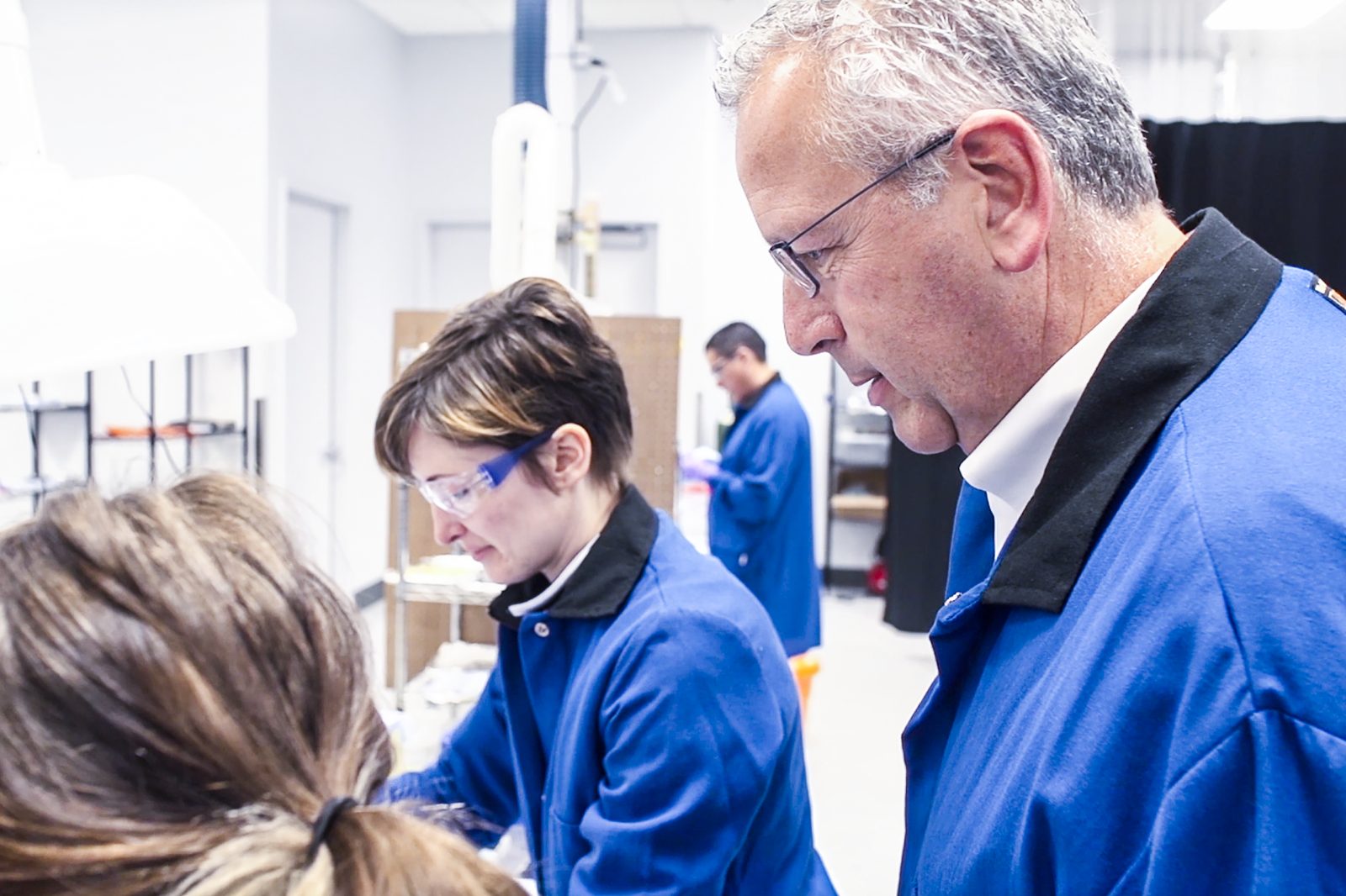 man and two women look at objects on a table both wearing blue lab coats