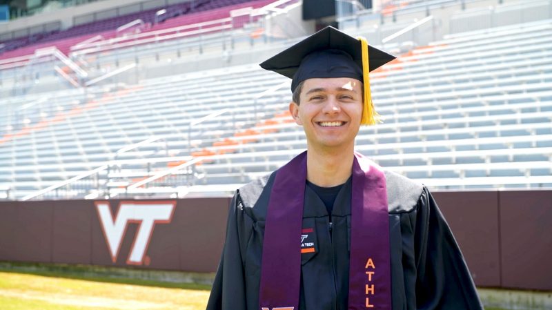 man wears graduation attire and stands in VT football stadium with empty stands