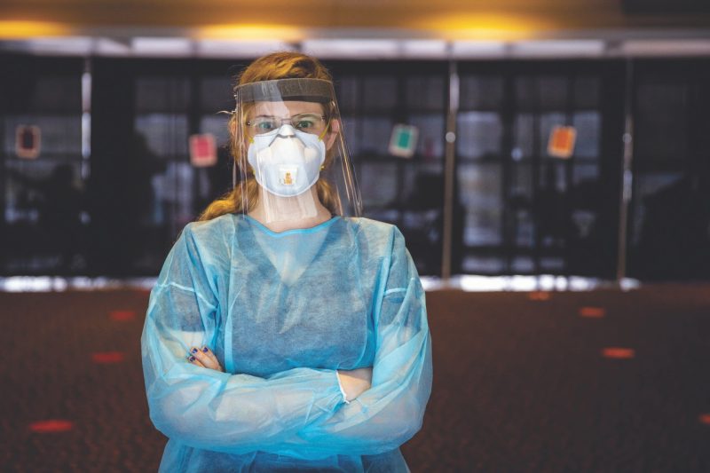 Vaughn stands in full protective gear (gown, mask, face shield) with arms crossed.