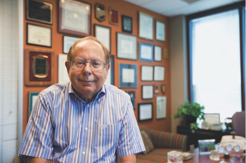 Montgomery sits on desk in office with a wall of awards and accolades in background.