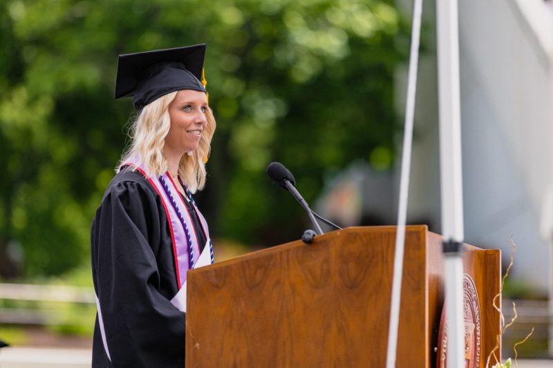 side view of woman in graduation attire speaking at a podium