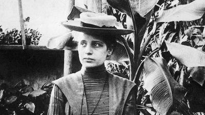 Woman wearing Victorian-era clothing and hat stands with hands crossed looking off in the distance in a room with plants and trees.