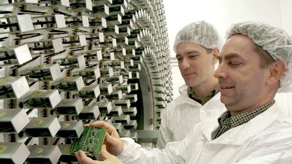 Two men wear lab coats and hairnets insert a cartridge into a circular machine.