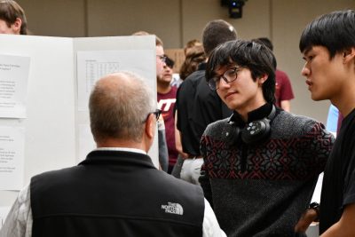 Man with back to camera talks to two students who are presenting a poster.