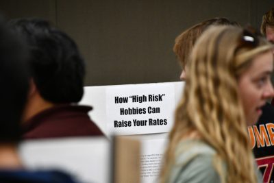 Poster reading "How High Risk Hobbies Can Raise Your Rates" is shown clearly in background, while students in foreground are blurred.