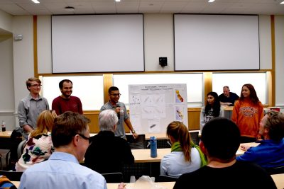 Five students stand next to poster board in front of classroom facing a group of judges.