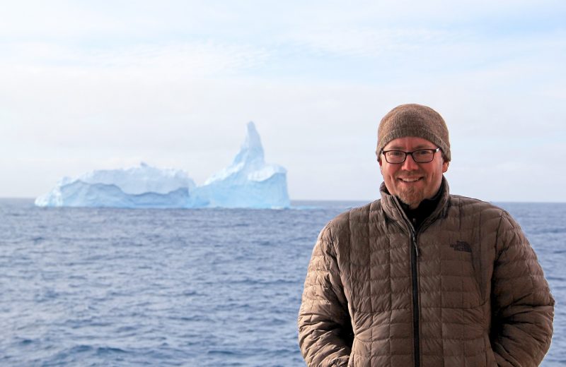 Brian Romans stands on the deck of the JOIDES, near West Antarctica, in January 2018.  Behind him is a massive iceberg in the distance. 