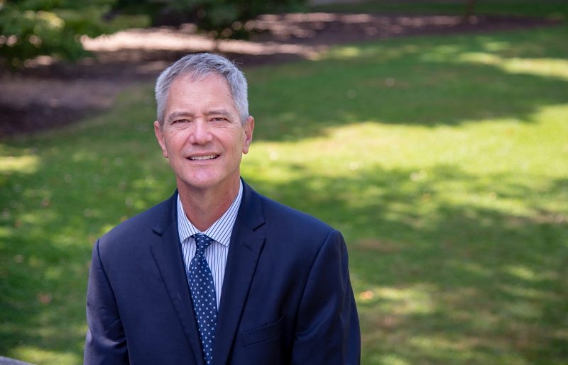 A man with salt and pepper hair wears a blue suit as he poses for a photograph. Behind him is bright green grass and the shadows of tall trees. 