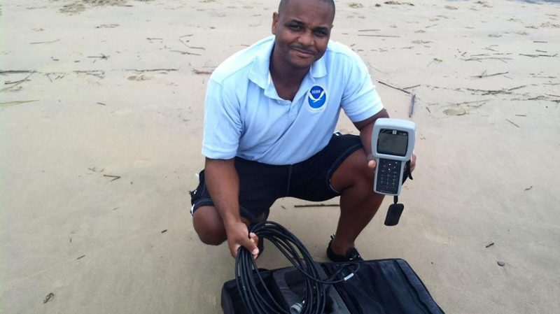 Scientist Lonnie Gonsalves squats on a beach, its sand wet, while holding wires and other pieces of hardware. 