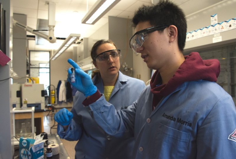 Amanda Morris in blue lab coat and gloves works with student Jie who looks at vial with purple powder