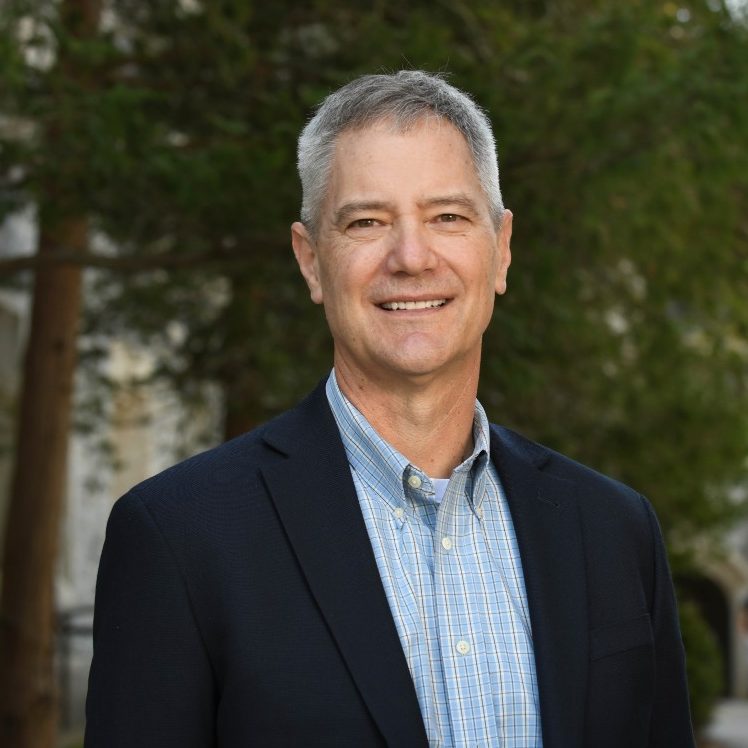 Pitts smiles outside with trees and stone buildings in background