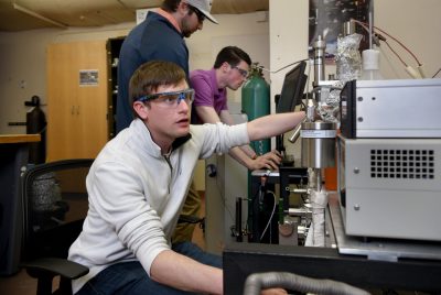 3 students wear lab googles and manipulate equipment in chemistry lab