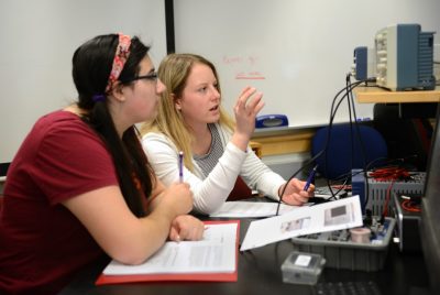 Two female students talk and look at device readout during physics lab.
