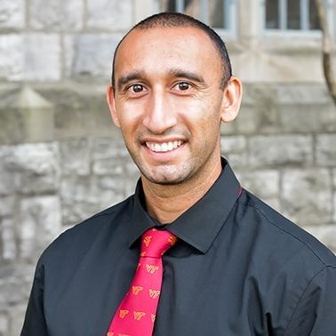 man in black shirt and Virginia Tech tie stands in front of stone wall and smiles at camera