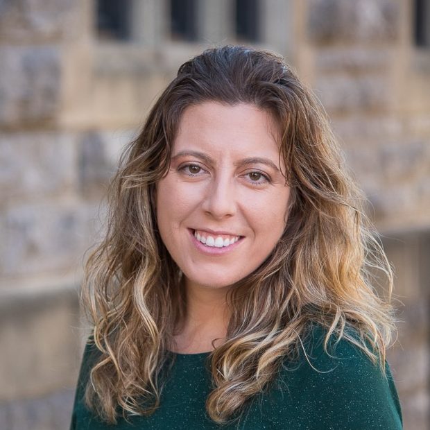 woman in dark green sweater stands in front of stone wall and smiles at camera