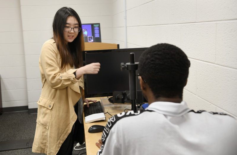 female student directs attention of male student looking through an eye tracking device.