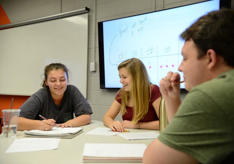 3 students sit at a table and work through math problems with pleasant discussion. Screen in background show the equation.