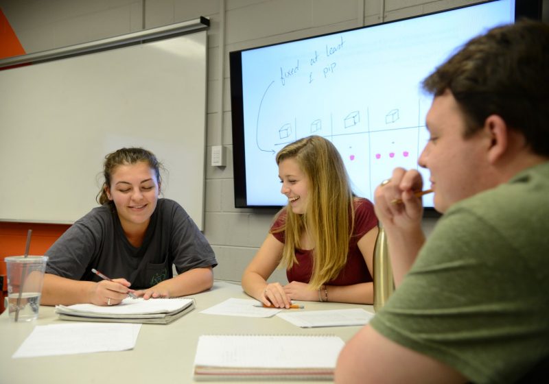 E Johnson Math Class (Spring 2019) 3 students sit at a table and work through math problems with pleasant discussion. Screen in background show the equation.