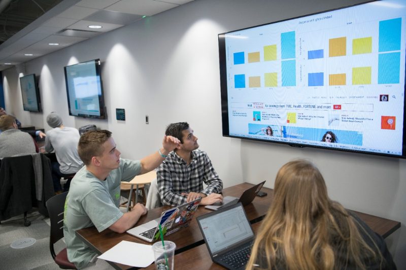 3 students analyze data and graphs on a wall display screen