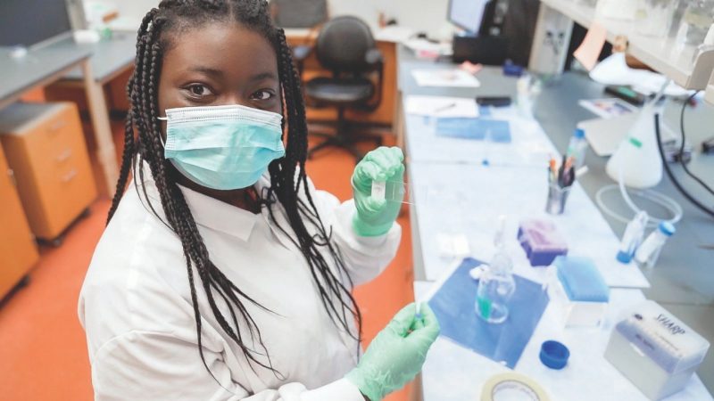 Niesha holds up a slide while seated at lab bench wearing lab coat, gloves, and mask.