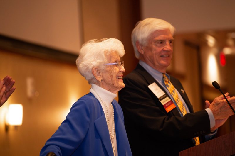 older female and male stand at podium and smile at audience