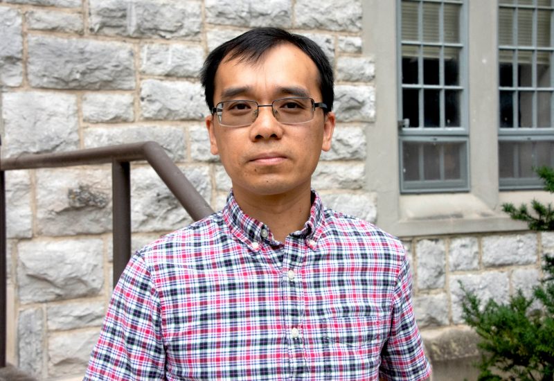 Xinwei in pink and blue plaid shirt outside with Hokie stone building in background