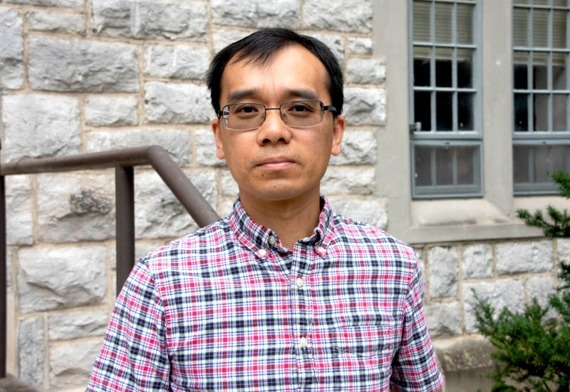 Xinwei in pink and blue plaid shirt outside with Hokie stone building in background