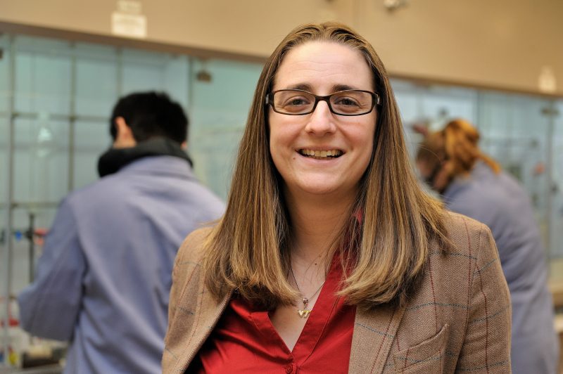 Amanda with red shirt and tan jacket stands in lab with students working in the background