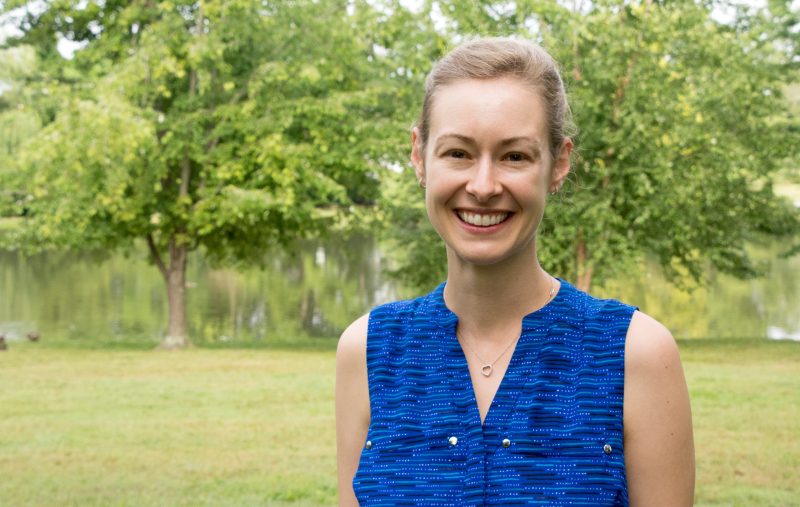 Cayelan in blue sleeveless shirt stands in front of tree-lined duck pond on sunny day