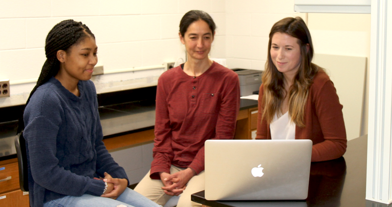 Langwig with 2 female students look at computer and smile