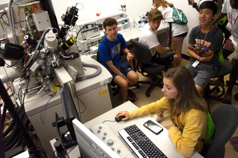 girl with yellow sweatshirt navigates computer with other students look a screen. Image taken from above.