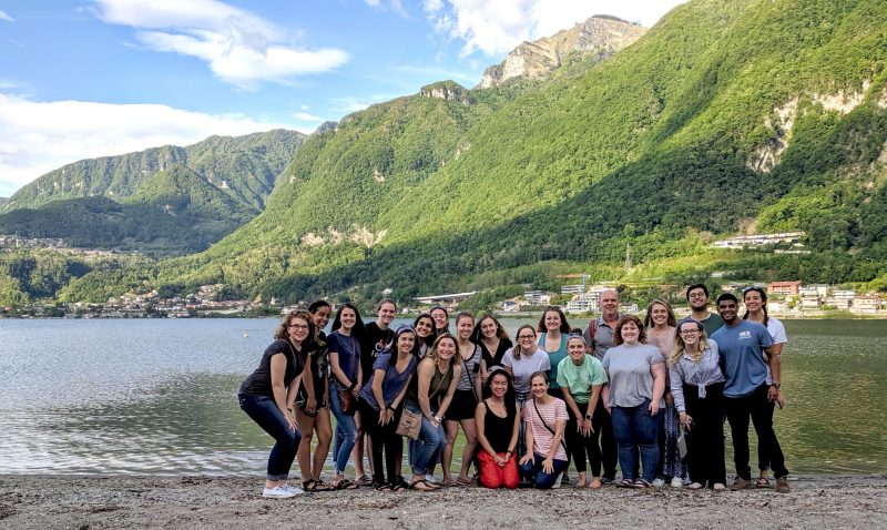 large group of neuroscience student's and director, Harry Sonteheimer, standing on a beach with water and Italian mountains in the background