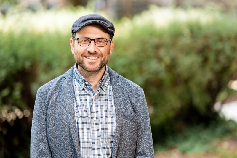 Serkan with news style cap and gray jacket stands outside with blurred greenery background