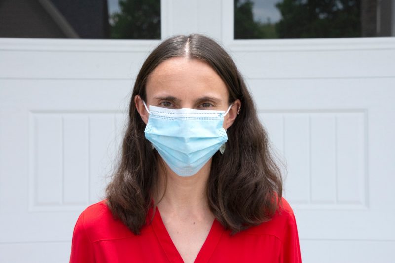 woman stands in front of door, looking at camera, wearing a mask