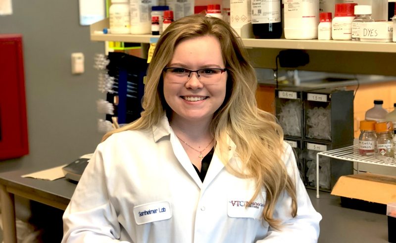 woman smiles and stands in front of lab bench, wearing a lab coat