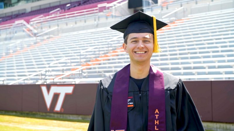 man wears graduation attire and stands in VT football stadium with empty stands