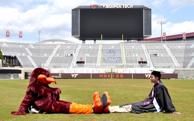 Hughes sits on football field in cap and gown across from Hokie Bird, feet to feet.