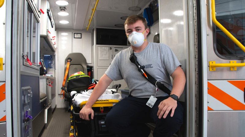 Buck wears a mask and kneels next to gurney inside ambulance.